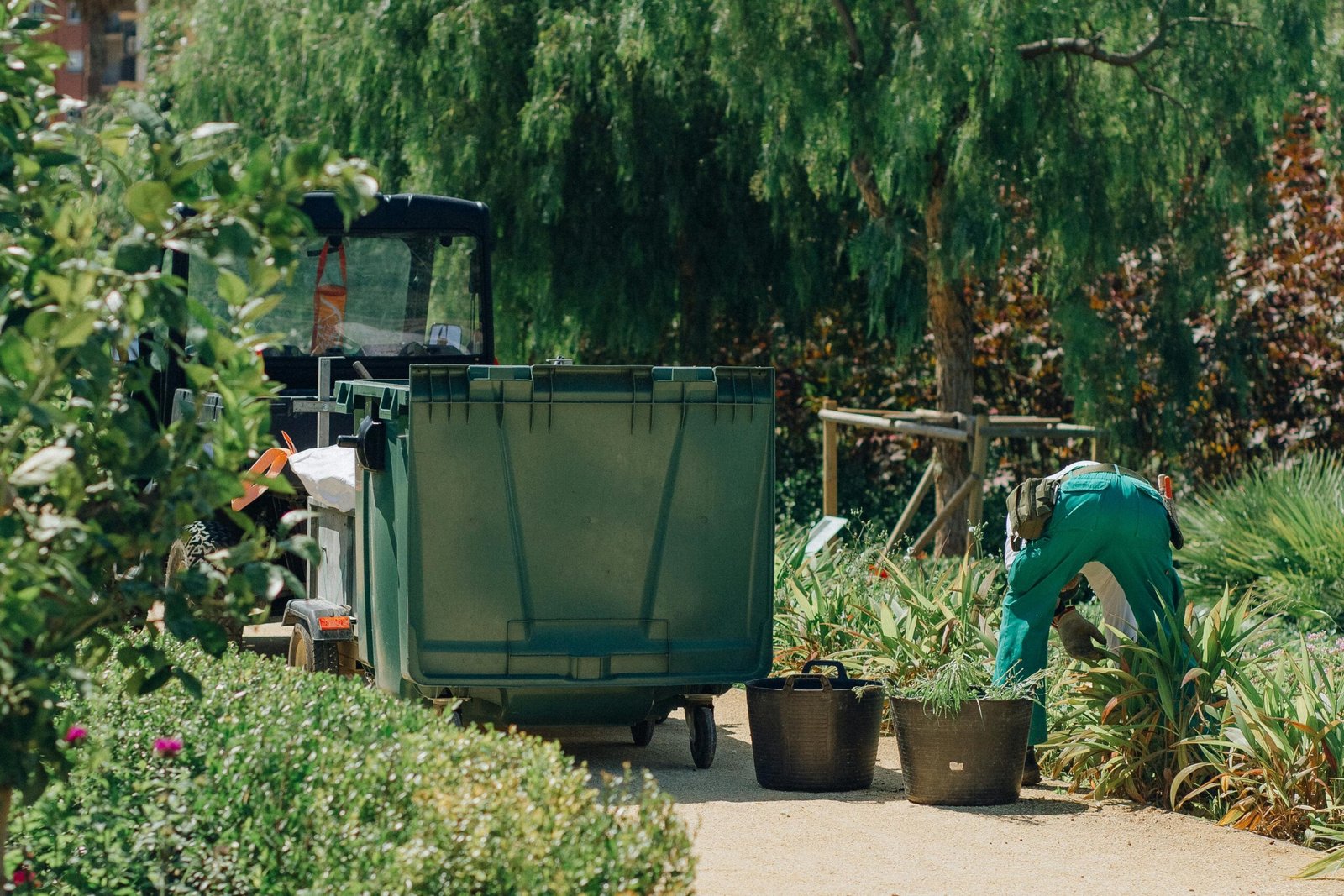 A gardener tends to plants beside a large green bin in a lush park environment.